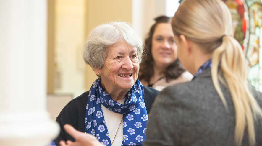 A woman with a short white bob wearing a blue forget-me-not scarf, smiles at a woman with long blonde hair she is in conversation with