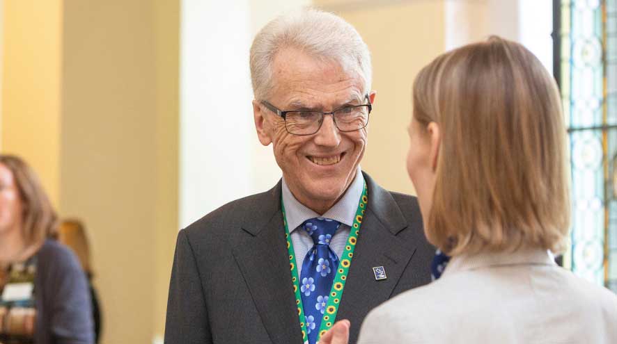 A man with white hair and glasses, wears a forget-me-not tie and a sunflower lanyard, smiles at a woman he is talking to - you only see the back of her head.