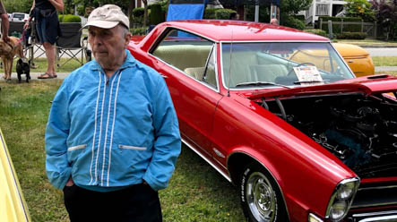 An older man in a blue jacket and baseball cap stands next to a red classic car