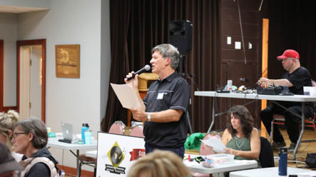 An older man stands holding a piece of paper and a microphone, speaking to a room of people
