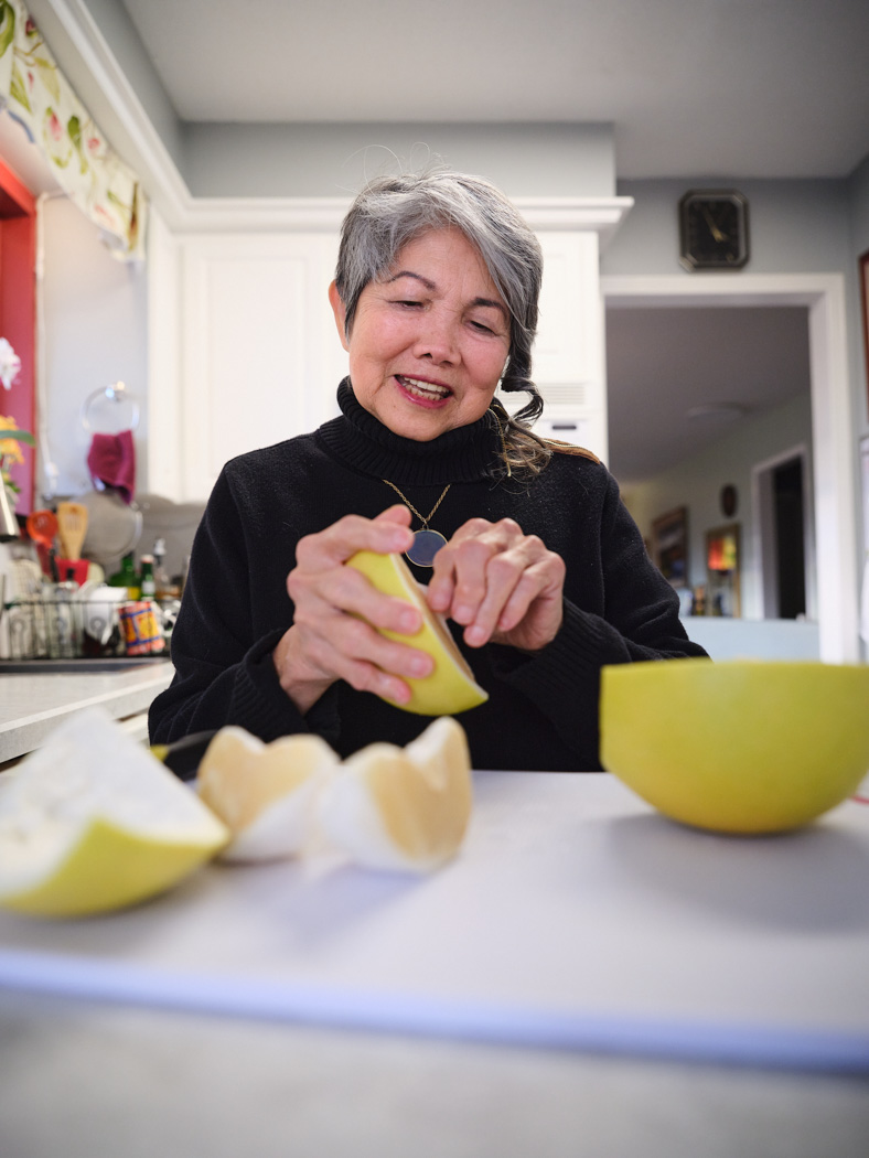 A woman smiles while peeling fruit at a kitchen counter, with apple pieces and a bowl in front of her.