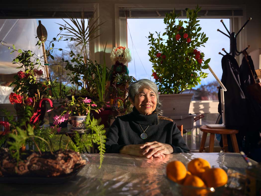 A woman sits at a table surrounded by colourful plants and flowers, with oranges in the foreground.