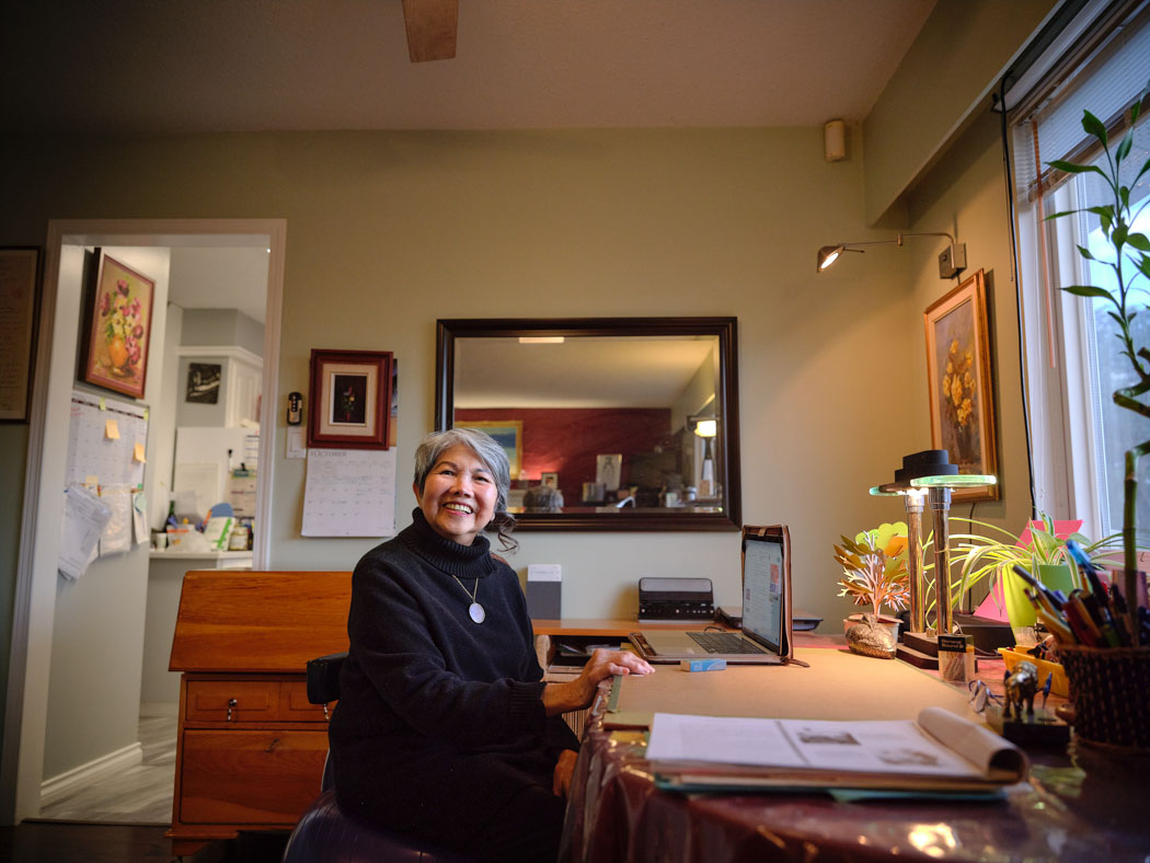 A woman sits at a desk in a bright home workspace surrounded by papers, artwork and plants.