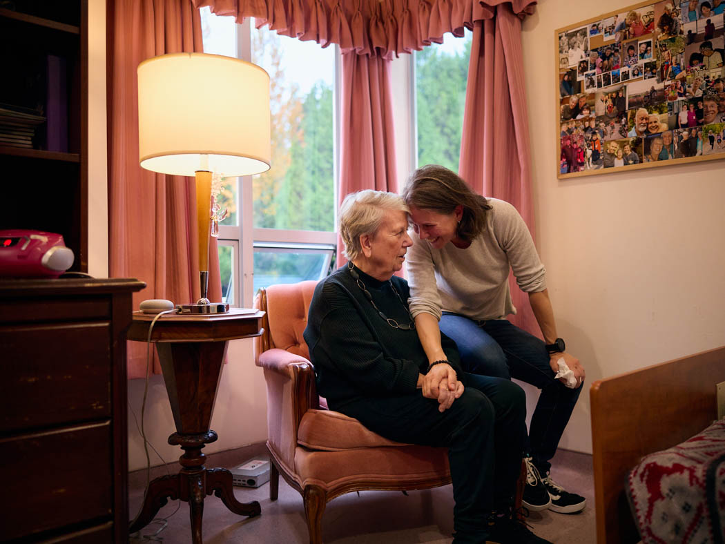 An older woman sits in an armchair while another woman leans in beside her, sharing a warm moment together.