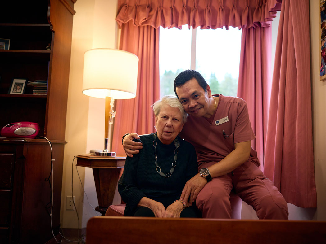 An older woman sits in an armchair while a man in scrubs sits close beside her with his arm around her shoulders.