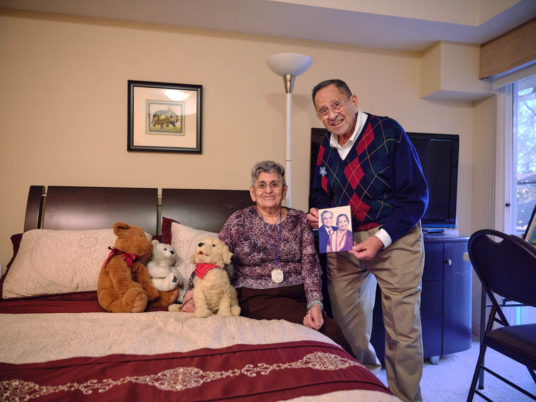 A woman sits on a bed beside several stuffed animals while a man stands next to her holding a framed photo.