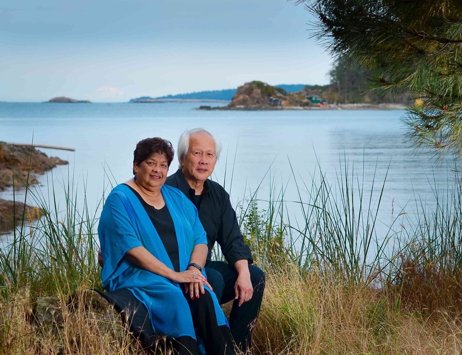 A woman sits next to her husband on a rock. The ocean is behind them.