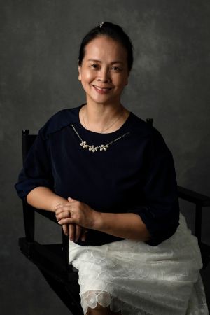 Portrait of Cynthia Wong, seated in a black wooden chair against a dark grey backdrop. She wears a navy blouse, delicate necklace and a white lace skirt, with her hands gently resting together.