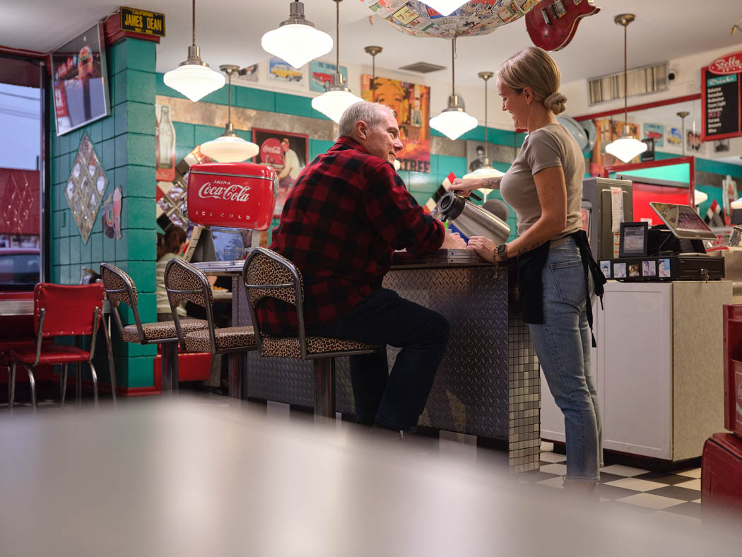 An older man sits at the counter of a colourful diner, talking with a staff member.