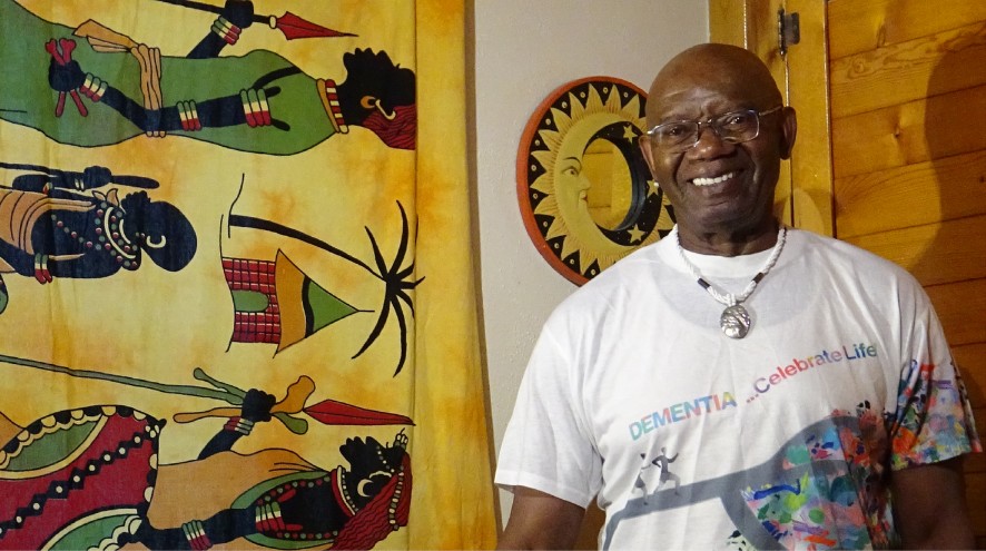 A man wearing glasses stands smiling in a room with African artwork on the wall.