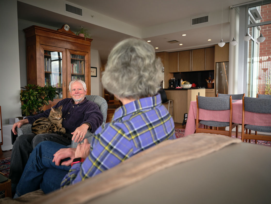 Two older adults sit facing each other in a comfortable shared living space, engaged in conversation.