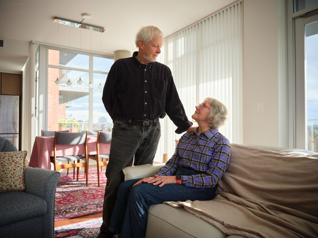 An older man stands beside an older woman seated on a couch as they smile at each other in a bright living room.