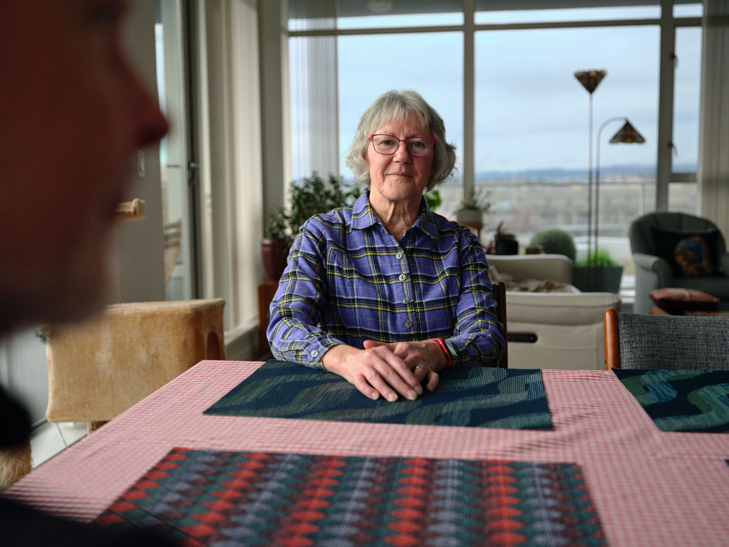 An older woman smiles while seated at a table, with another person in the foreground and a large window overlooking the city behind her.