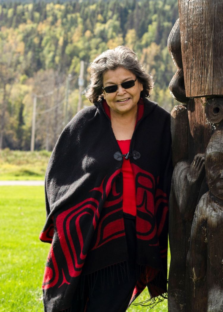 An Indigenous Elder leans against a totem pole, smiling, surrounded by grass and trees.