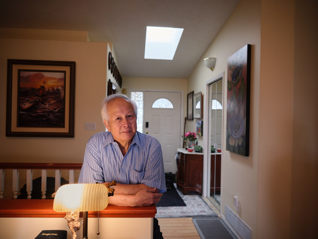 An older man leans over a banister with his arms folded in a bright hallway of his home.