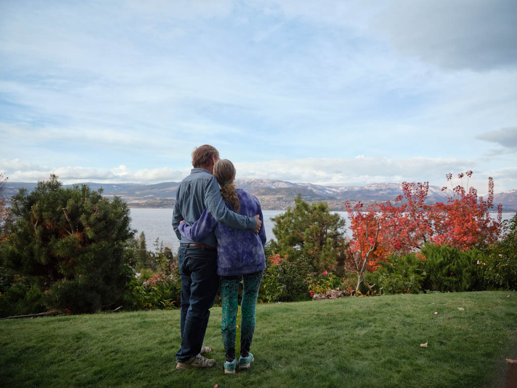An older couple stand with their arms around each other, looking out over a scenic landscape with autumn foliage.
