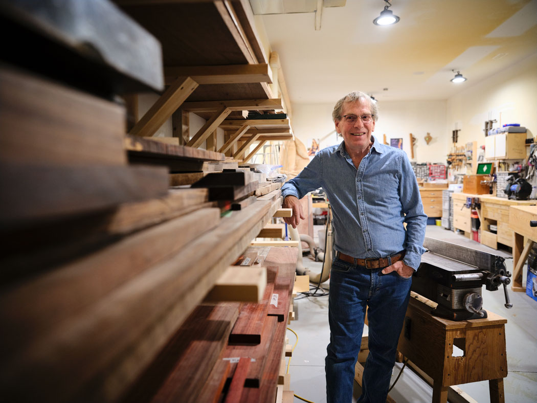 An older man stands beside a workbench in a woodworking shop, surrounded by tools and lumber.