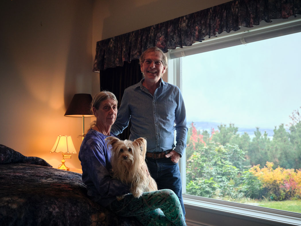 An older couple pose with a small dog beside a large window overlooking a green landscape.