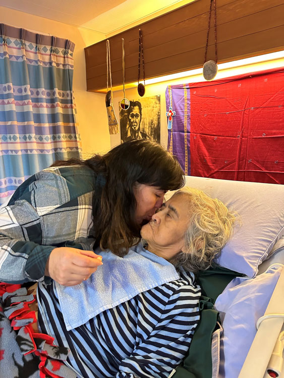 A woman leans over to kiss an older woman resting in bed.