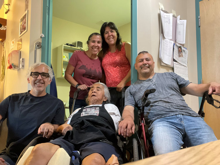 Four adults gather around an older woman seated in a chair, smiling together.