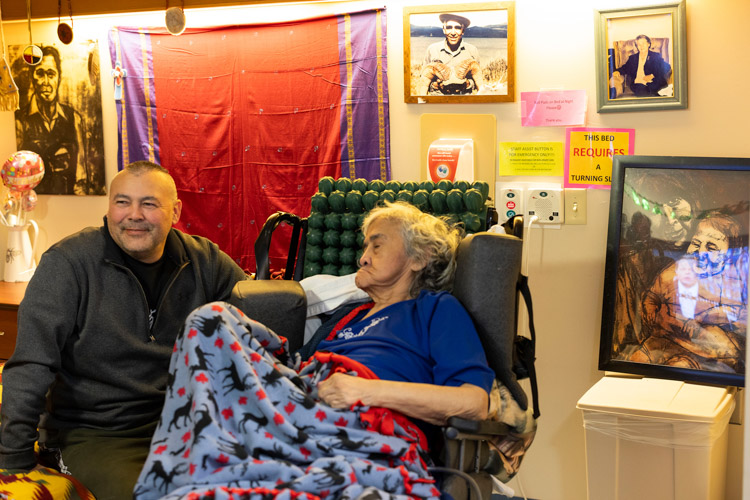 An older woman sits wrapped in a colourful blanket beside a smiling man, with family photos and a framed portrait displayed around them.