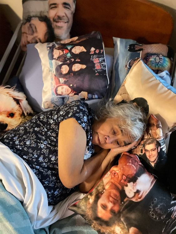 An older woman rests in bed surrounded by pillows printed with photos of loved ones.