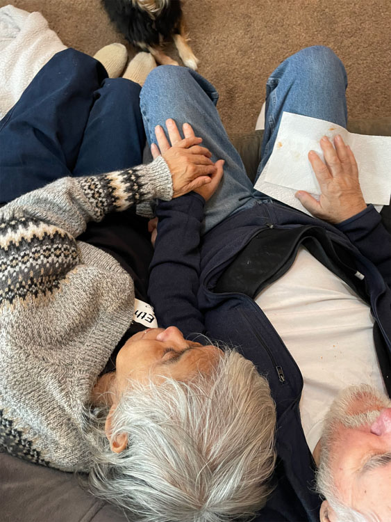 An older couple lie side by side, holding hands while reading together.