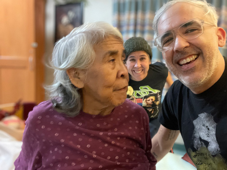 An older woman sits beside a smiling man, with another person smiling in the background.
