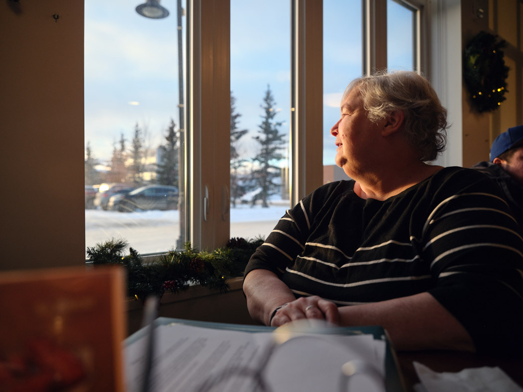An older woman sits at a table looking out a large window at a snowy landscape.