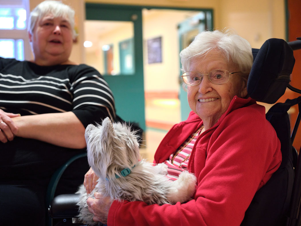 An older woman smiles while holding a small dog on her lap, with another woman seated beside her.