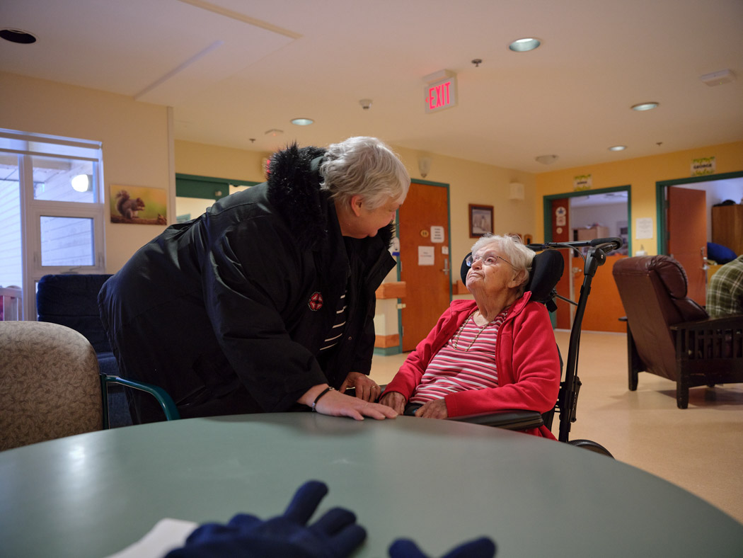 An older woman leans in to talk with another older woman seated in a wheelchair in a shared living space.