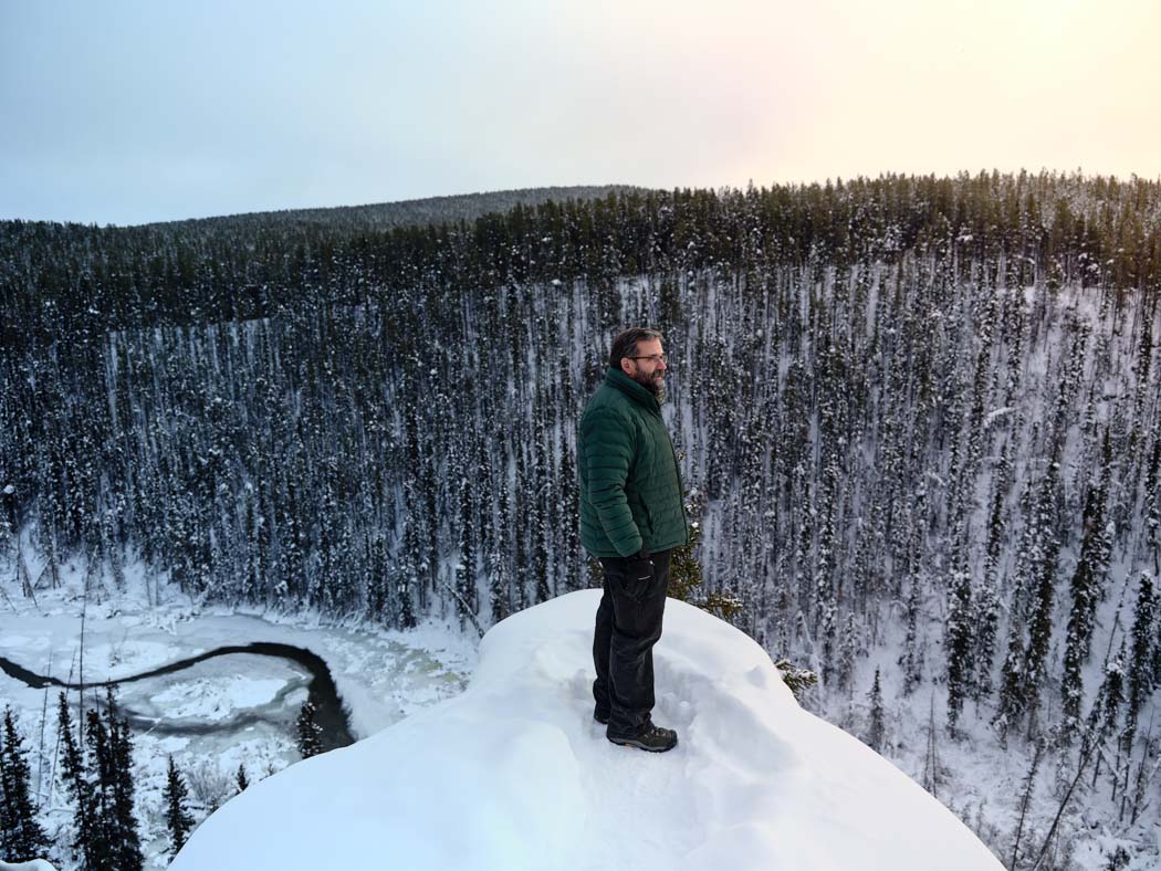 A person stands on a snowy overlook surrounded by a vast, snow-covered forest.