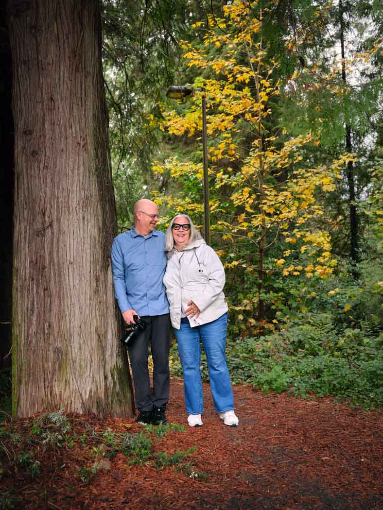 Two people stand close together, smiling, beside a large tree in a wooded area with autumn foliage.
