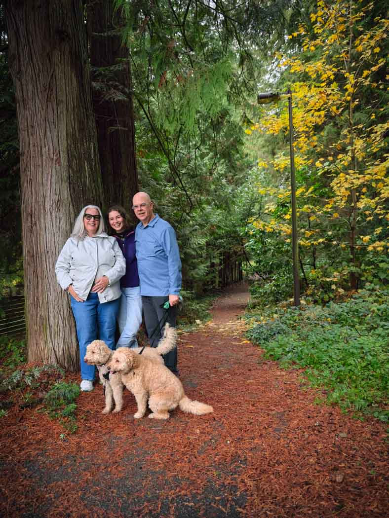 Three people stand close together, smiling, with two dogs beside a large tree on a wooded path surrounded by autumn foliage.