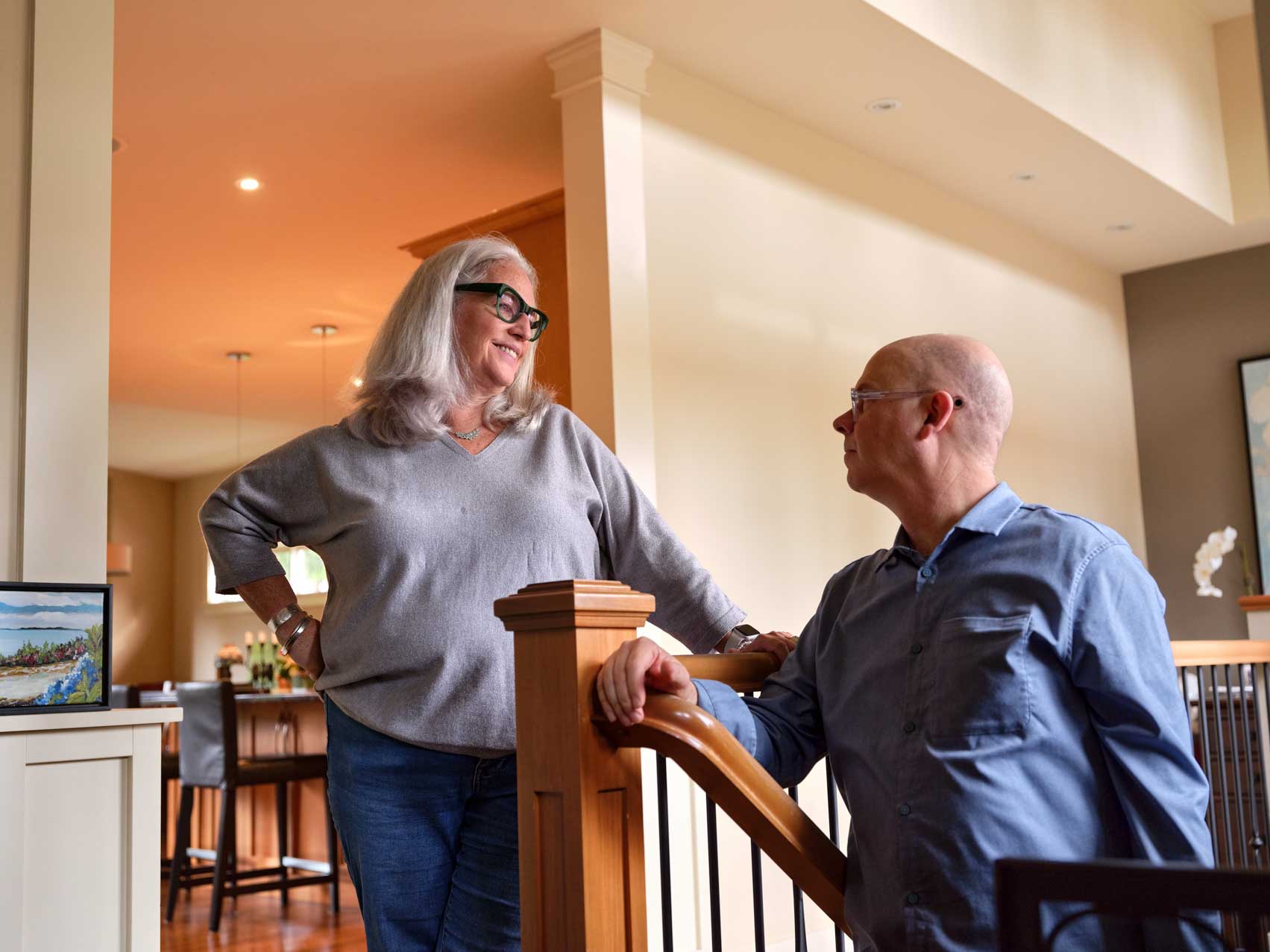 A woman stands beside a staircase talking with a seated man in a bright, open home.
