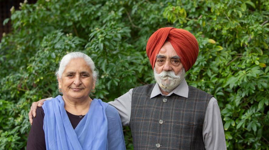 An elderly couple with the husband putting his arm around his wife, posing together in front of trees.