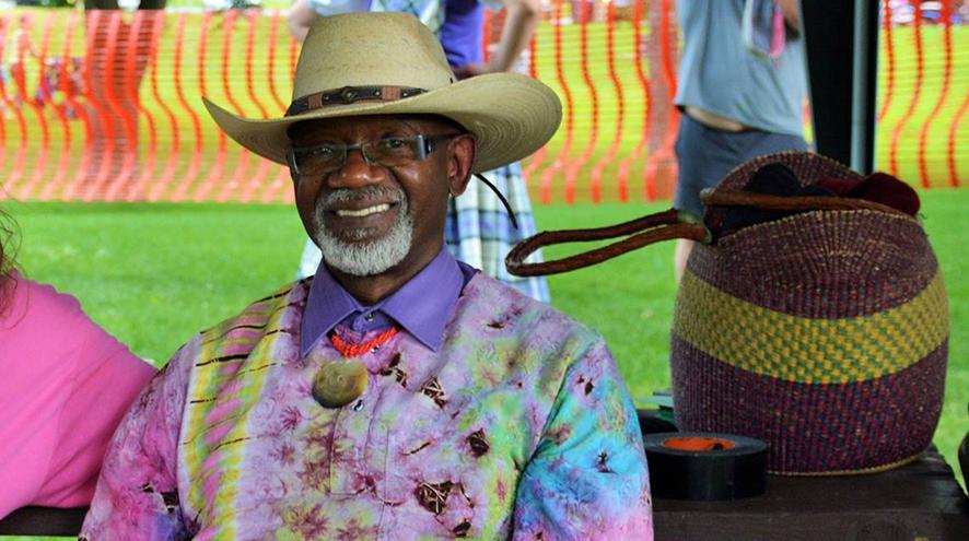A black man with a short grey beard sits on a bench during an outdoor festival, wearing a wide-brimmed hat.