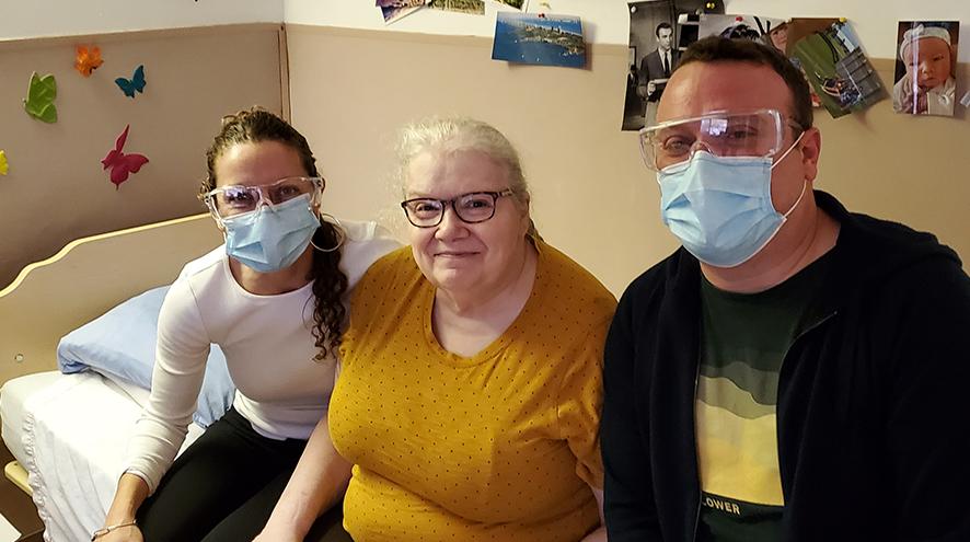 A young woman and a young wear surgical facemasks on either side of a shorter, older woman on a bed.