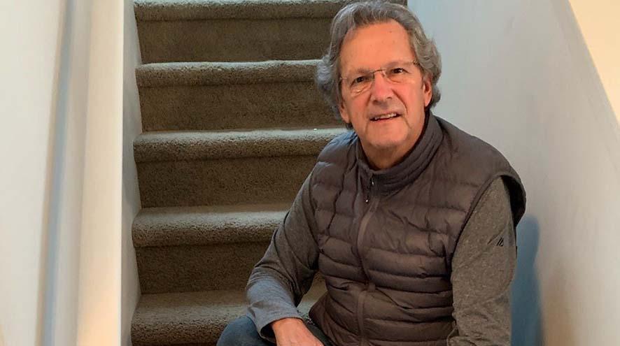 An older man with wavy grey hair sits on a set of indoor stairs, wearing a puffy vest.