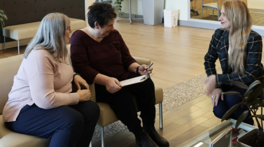 Two women in a bank, sitting in the lounge, speaking to an employee