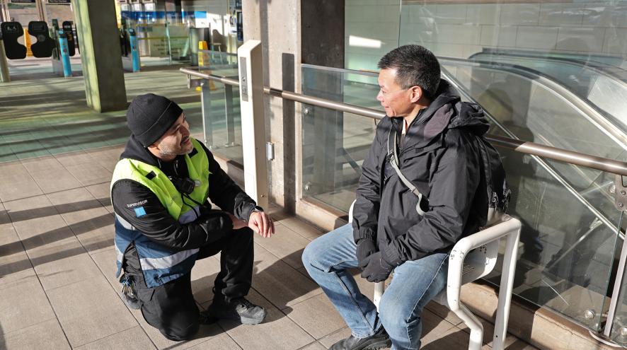 A transic police officer kneels in front of a man on a bench. He is making eye contact, and offering to help. 