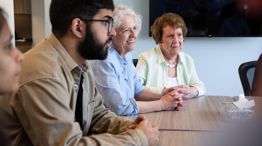 Three women and one man of mixed ages sit on one side of a wooden table, looking at someone on the other side with supportive expressions.