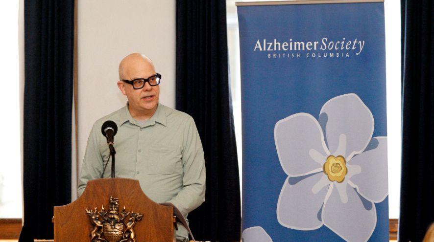 A man, Geoffrey, wearing thick black glasses addresses a crowd from a podium next to a blue banner with a forget-me-not-flower