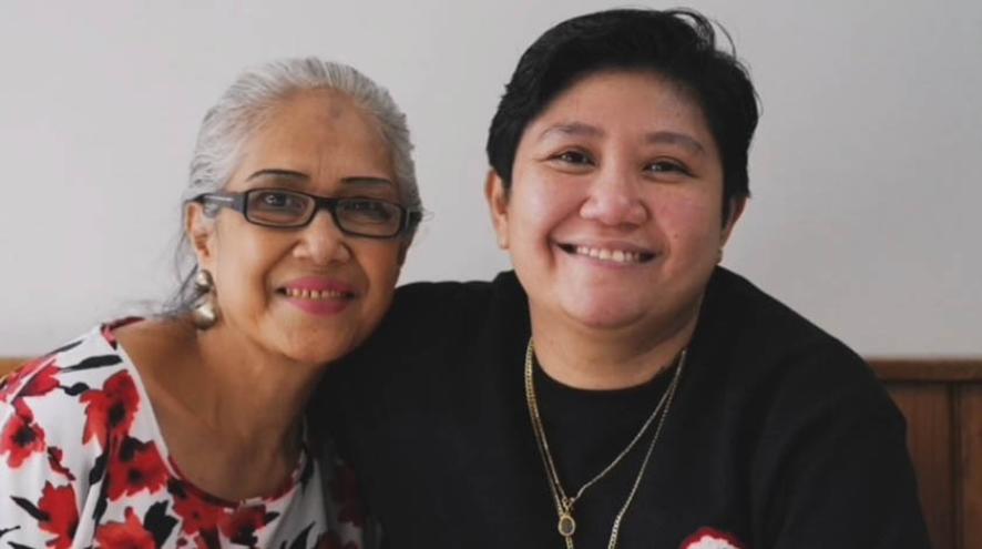 An older woman with gray hair and glasses, smiles and poses close to a younger person with short dark hair