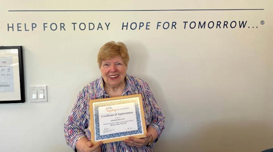 Happy older woman stands in front of a white office wall holding a certificate of recognition 