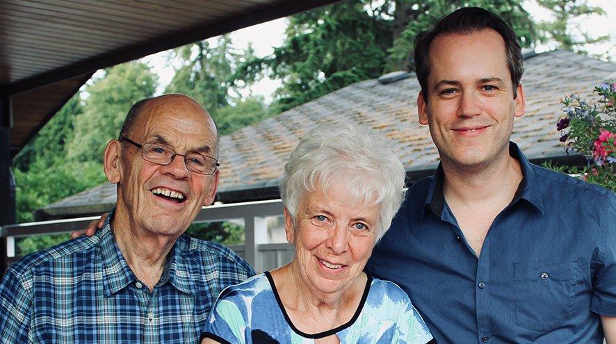 An elderly couple stands outside with their adult son. They are all wearing blue shirts and smiling.