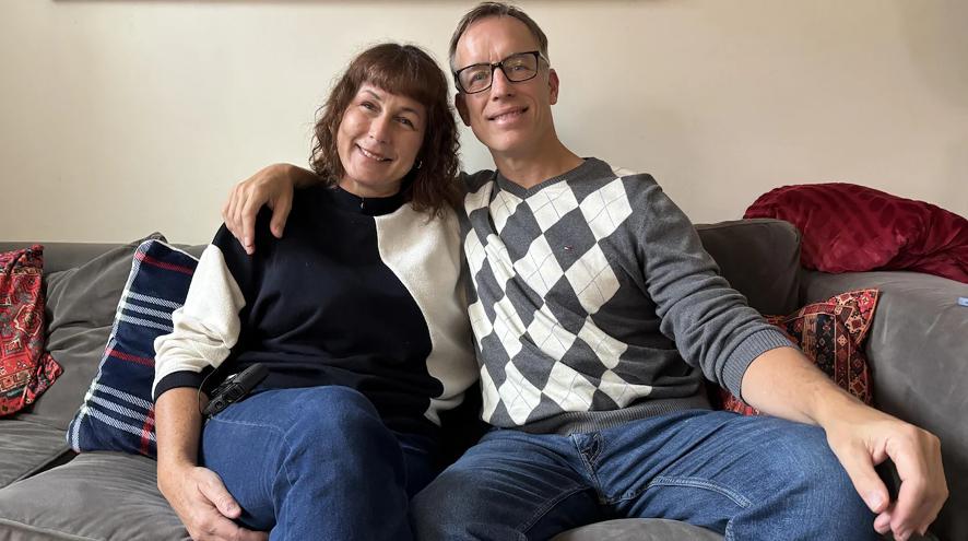 Karina and Andrew Reid are sitting next to each other on their living room couch, with Andrew's arm on Karina's shoulder.