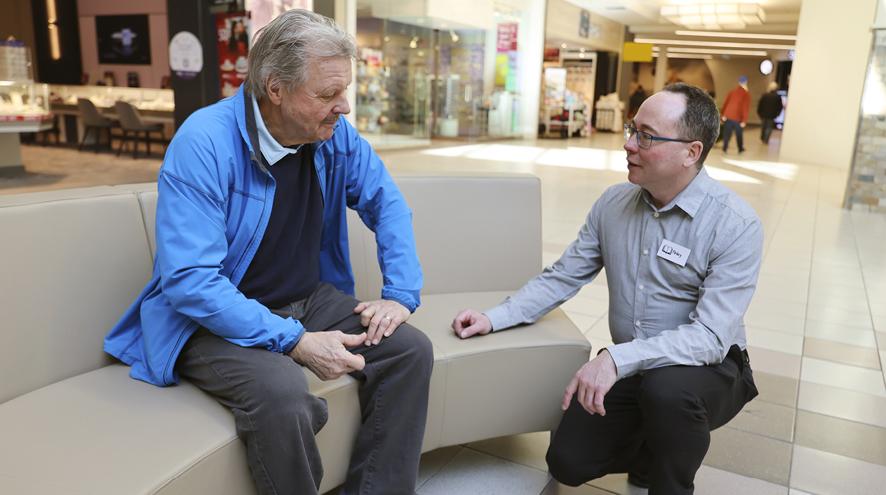 Inside a shopping mall, a gentleman is sitting on a bench while talking to a worker. The worker is kneeling, speaking to the man at eye level.