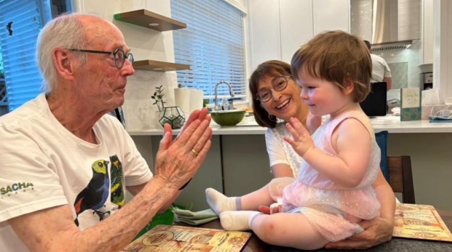 Lived experience Dan is playfully clapping his hands in front of his grand draughter, who is sitting on the dining table and held by her grandmother.