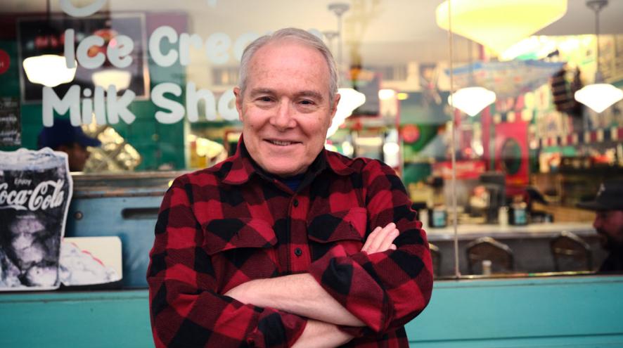 An older man in a red-and-black plaid shirt smiles with his arms crossed inside a colourful diner.
